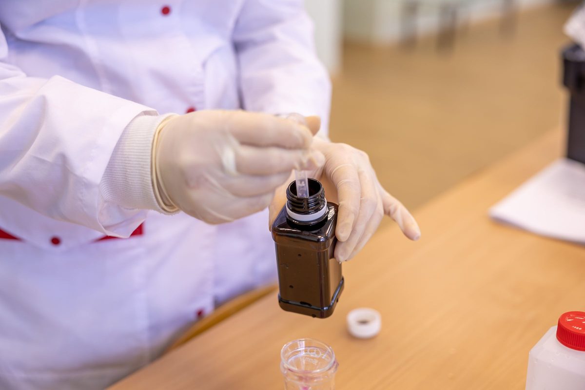 Gloved hands drawing a sample from a brown lab bottle with a pipette